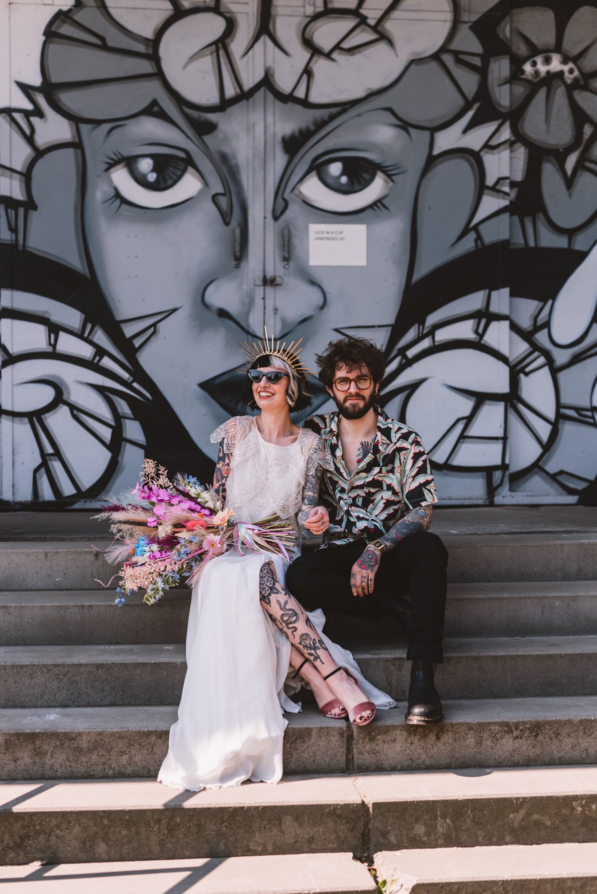 Couple seated on step in front of graffiti wall holding bouquet
