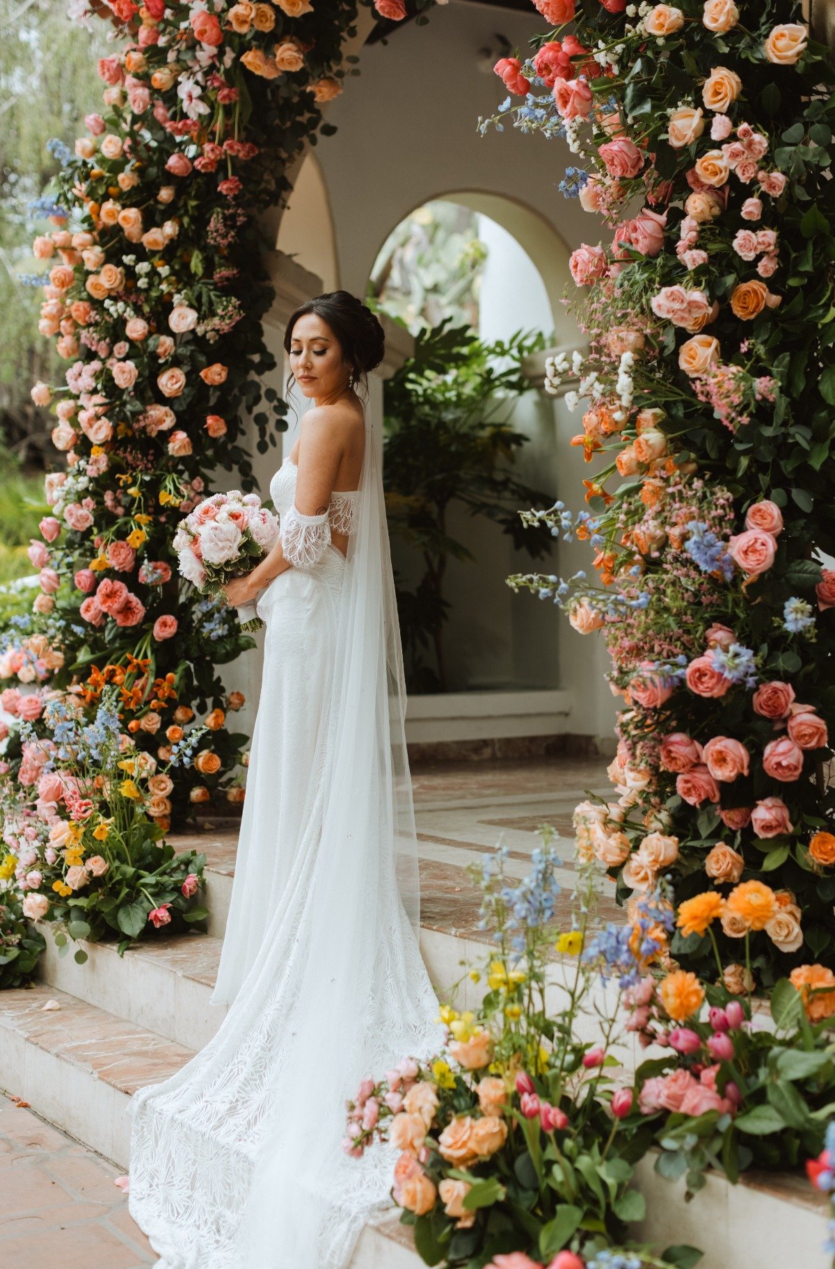A bride in a white dress is standing under a floral archway.