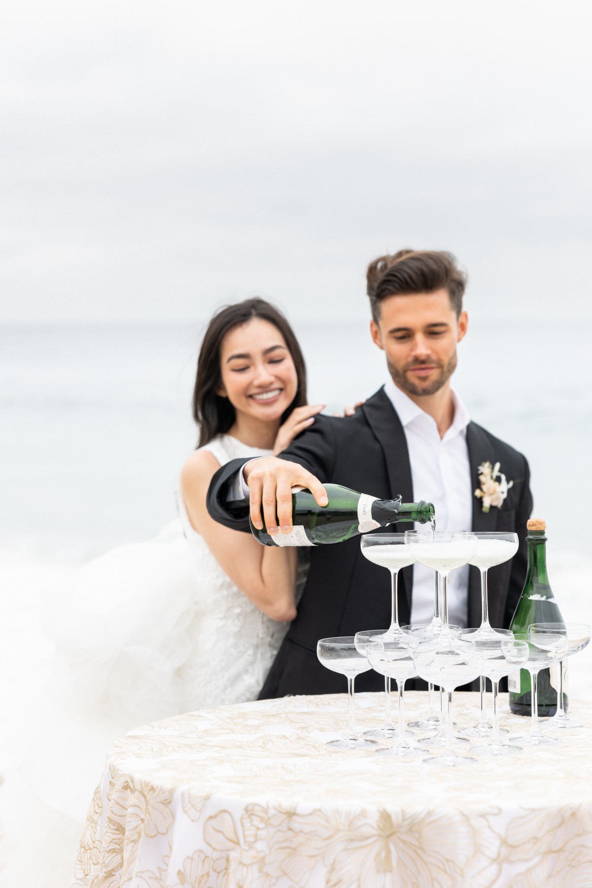 Bride and groom pouring timeless champagne tower at beach