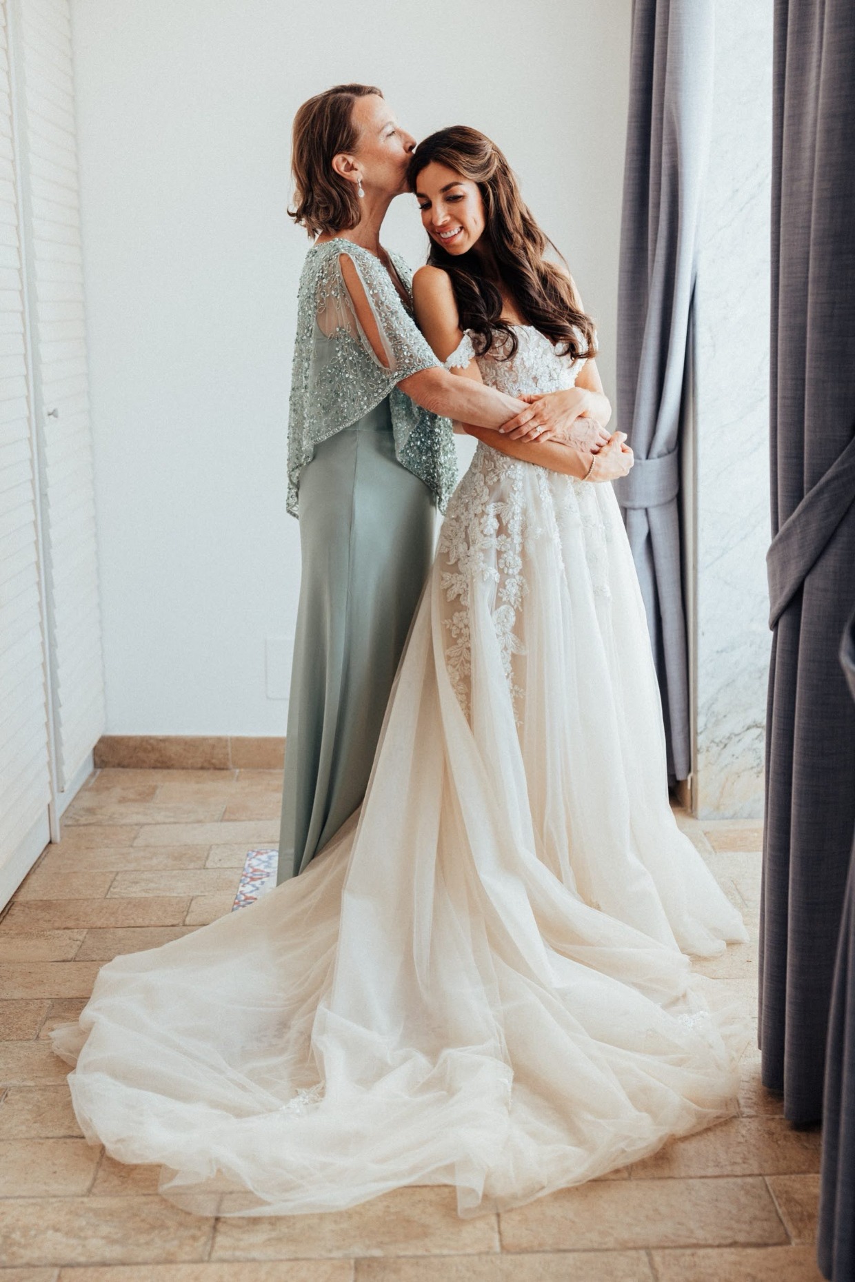 Mom kissing bride's head while helping her get ready in dress