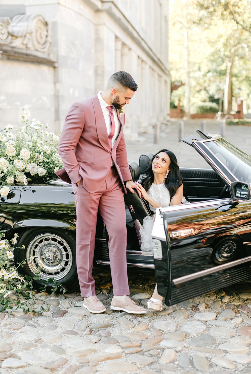 groom in mauve suit with bride in vintage car