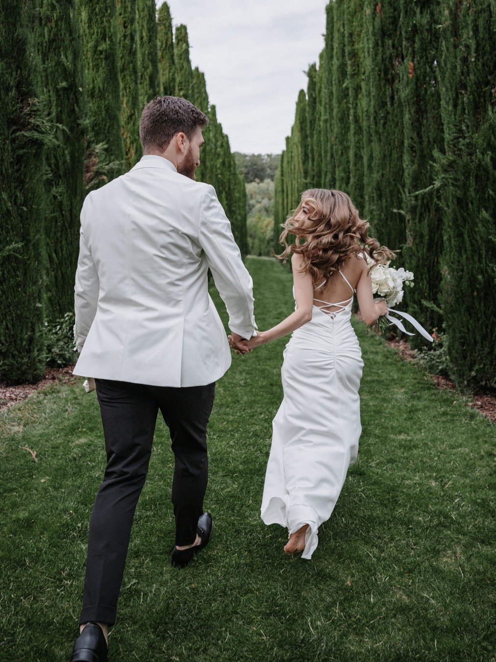 bride and groom running through a cypress tree garden